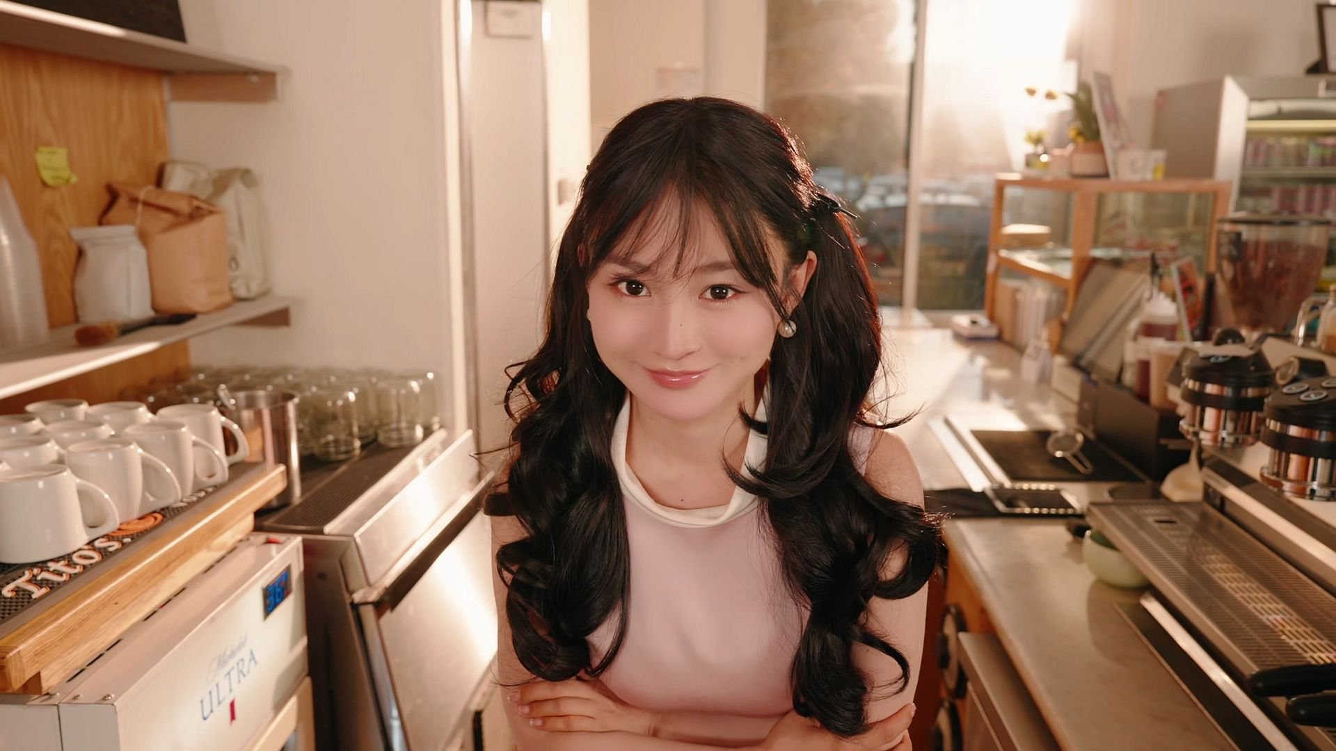 With her hair in twin pigtails, LI Sisi has her arms folded across her chest and looks at you inside a kitchen setting, with cups lined along cutting boards stacked to her left.