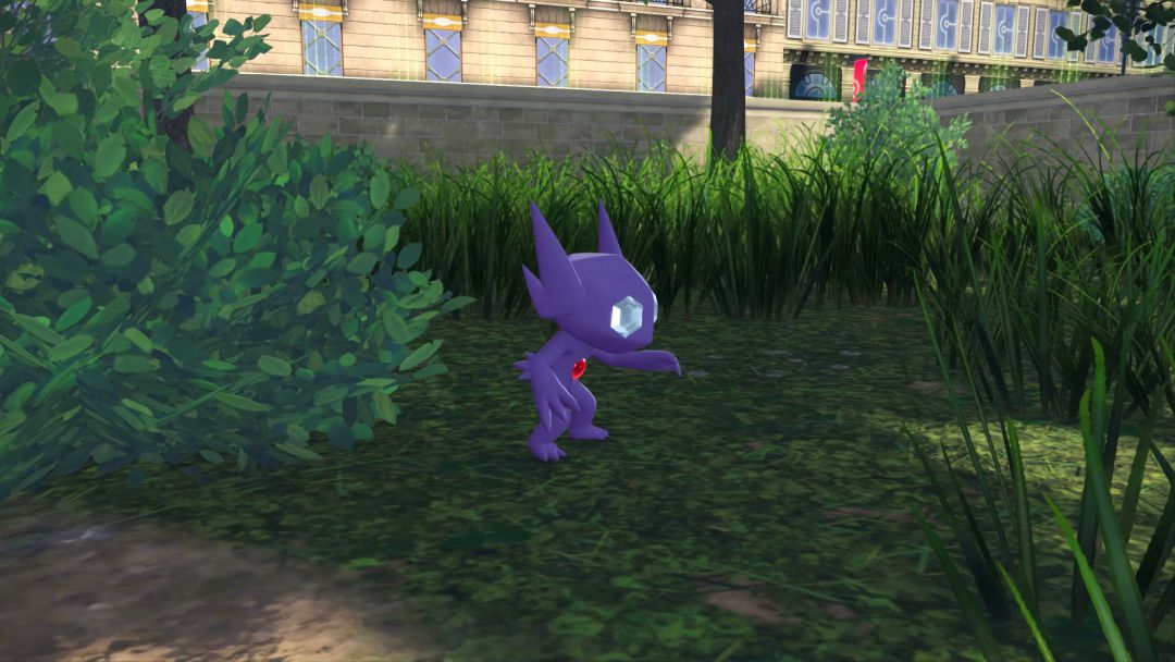 A closeup as Sableye emerges from some brush at the edge of a clearing, with grass, tree trunks, and a stone wall behind it.